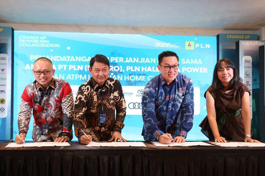 Four individuals, three men and one woman, are standing side by side, smiling, and signing documents on a table. They are dressed in colorful, patterned batik shirts. Behind them is a large screen displaying the logos of BUMN and PLN, along with a message about a partnership agreement.