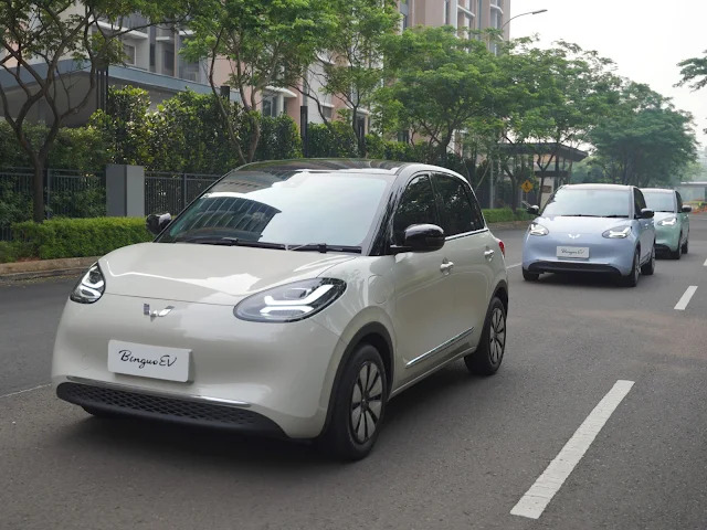 Sebuah mobil BinguoEV putih memimpin deretan kendaraan listrik sejenis yang melaju di jalan kota yang dipenuhi pohon dan bangunan di latar belakang.